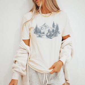 May include: Cream-colored t-shirt featuring a mountain and forest landscape illustration in shades of blue. The graphic includes a waterfall, trees, and a mountain range. The shirt has short sleeves and a crew neck.
