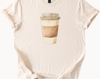 Puede incluir: Camiseta color crema con una ilustración en acuarela de una taza de café con tapa. La taza tiene una manga marrón y una tapa marrón claro. La camiseta es de material suave y tiene cuello redondo.
