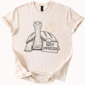 May include: A cream-colored short-sleeve t-shirt featuring a dark gray graphic of a turtle with a long neck and a grumpy expression. The turtle holds a sign that reads NOT IMPRESSED, with two small yellow starburst designs above its shell.