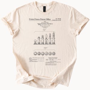 May include: Cream-colored t-shirt featuring a black and white patent diagram of chess pieces. The diagram includes detailed illustrations of each piece and the text "United States Patent Office". The shirt has a classic crew neck and short sleeves.