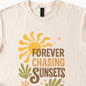 May include: Cream-colored t-shirt featuring a retro design with a yellow sun, green and orange foliage, and the text "FOREVER CHASING SUNSETS" in brown and orange. The shirt has a crew neck.