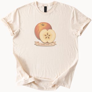May include: A cream-colored t-shirt featuring a graphic of a whole red apple and a sliced apple half. Below the fruit is a tan banner with the text "COX'S ORANGE PIPPIN - EST.". The shirt has short sleeves and a crew neckline, presented in a flat view.