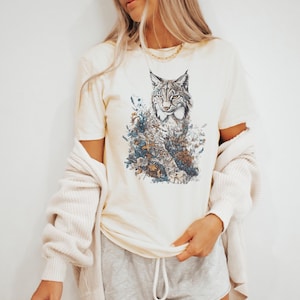 May include: Cream-colored t-shirt featuring a detailed illustration of a lynx surrounded by floral elements in shades of blue, brown, and green. The short-sleeved tee is made of soft material. A light beige cardigan is draped over the shoulders.