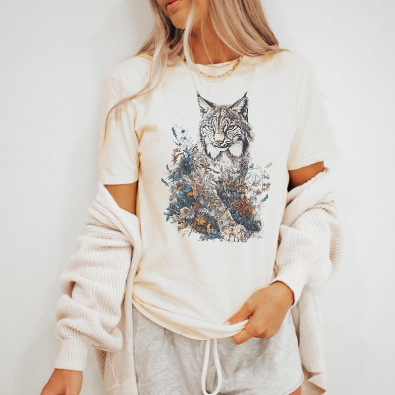 May include: Cream-colored t-shirt featuring a detailed illustration of a lynx surrounded by floral elements in shades of blue, brown, and green. The short-sleeved tee is made of soft material. A light beige cardigan is draped over the shoulders.