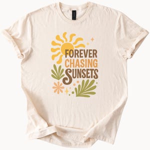 May include: Cream-colored t-shirt featuring a retro design with a yellow sun, green leaves, and the text "FOREVER CHASING SUNSETS" in brown and orange. The shirt has short sleeves and a crew neck.