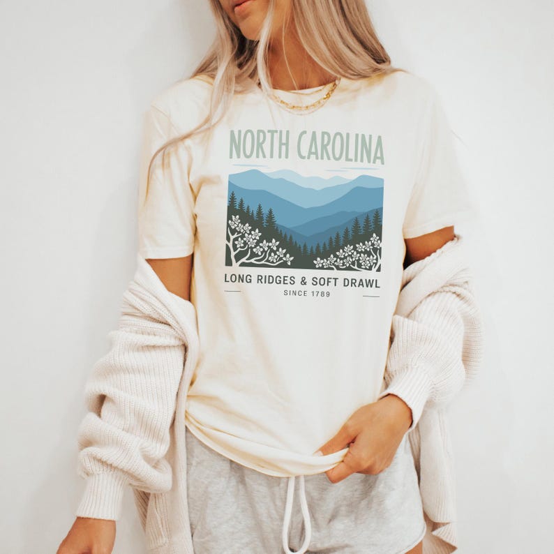 May include: Cream-colored t-shirt featuring a graphic of North Carolina mountains in shades of blue and green. The text "NORTH CAROLINA" is above the graphic, with "LONG RIDGES & SOFT DRAWL SINCE 1789" below. The shirt has short sleeves.