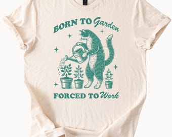May include: Cream-colored t-shirt with a teal graphic. The graphic features a cat watering potted plants with a watering can, with the text "BORN TO Garden" above and "FORCED TO Work" below. The shirt has short sleeves and a crew neck.