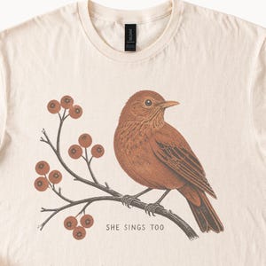 May include: A cream-colored t-shirt featuring a brown bird perched on a branch with red berries. The graphic includes the text "SHE SINGS TOO" printed in a simple font below the branch. The shirt has a crew neckline and a black brand label at the neck.
