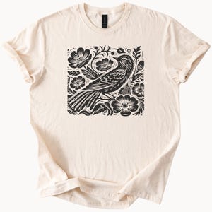 May include: A cream-colored t-shirt with a black and white graphic print of a bird surrounded by flowers.