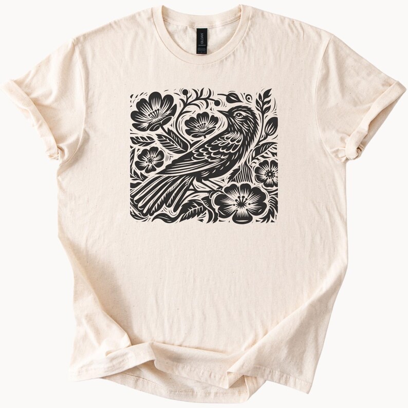 May include: A cream-colored t-shirt with a black and white graphic print of a bird surrounded by flowers.