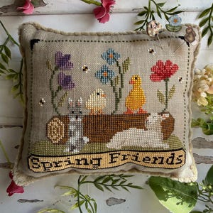 May include: A beige, rectangular, cross-stitched pillow featuring a spring theme. The design includes a bunny, two chicks, and a duckling, with flowers and the text "Spring Friends". The pillow has a fringed edge and is surrounded by floral accents.
