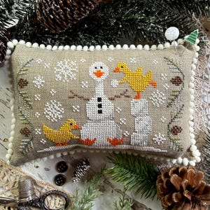 May include: A rectangular beige fabric pincushion with white pom-pom trim featuring a cross-stitch design of a white snowman, two yellow ducks, and white snowflakes. The design includes brown stick arms, orange feet, and green pine branch accents.