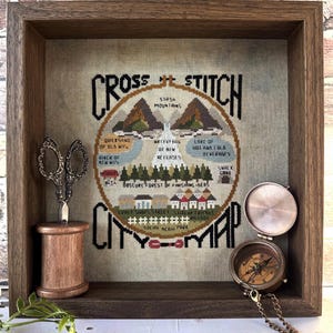 Può includere: Una mappa a punto croce incorniciata in una cornice di legno marrone. La mappa presenta un design circolare con testo e illustrazioni di montagne, una cascata e edifici. Un paio di forbici, una bobina di legno e una bussola sono posizionati vicino alla mappa.