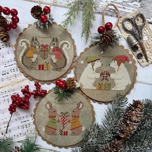 May include: Three circular cross-stitch ornaments on linen fabric featuring pairs of squirrels, polar bears, and rabbits exchanging gifts. Each ornament is topped with a pinecone, red berries, and a jute twine loop for hanging.