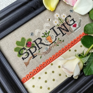Può includere: Un ricamo a punto croce incorniciato in legno nero, che presenta la parola "SPRING" in un carattere fantasioso con un coniglio, fiori e un annaffiatoio. La cornice è decorata con un tessuto bianco con pois verdi e un bordo arancione a festoni.