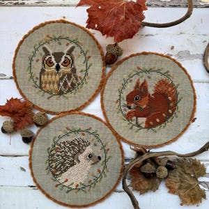 May include: Three circular cross-stitch designs: an owl, a squirrel, and a hedgehog, each encircled by a wreath of leaves and berries. They have a beige background with a brown, fluffy border. Acorns and autumn leaves are scattered around the designs.