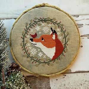 May include: A round, cross-stitched embroidery featuring a fox with orange and white fur, a red bird, and snowflakes. The fox is surrounded by a green wreath with red berries. The embroidery is set in a round frame with a light green fabric.