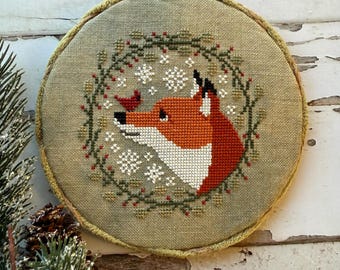 May include: A round, cross-stitched embroidery featuring a fox with orange and white fur, a red bird, and snowflakes. The fox is surrounded by a green wreath with red berries. The embroidery is set in a round frame with a light green fabric.