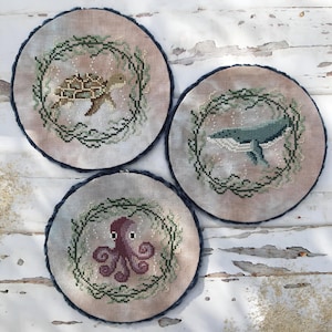 Può includere: Tre ricami a punto croce rotondi raffiguranti creature marine: una tartaruga, una balena e un polpo. Ogni disegno è circondato da una ghirlanda verde e posto su uno sfondo neutro. I bordi sono rifiniti con un bordo blu scuro.