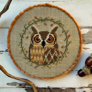 May include: A cross-stitch owl design in a circular frame. The owl is brown and tan, with large orange eyes, and is surrounded by a green and orange floral wreath. The frame has a tan fabric centre and an orange, fuzzy border.