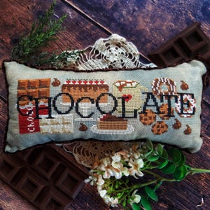 May include: A rectangular, cross-stitched pillow with the word "CHOCOLATE" and images of chocolate desserts. The pillow is light blue with brown trim and is surrounded by chocolate bars, flowers, and lace on a wooden surface.