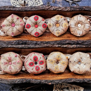 May include: Six round pincushions with different floral and geometric designs. The pincushions are made of fabric and have a beige background with colorful embroidery. They are arranged in two rows of three on a wooden surface.