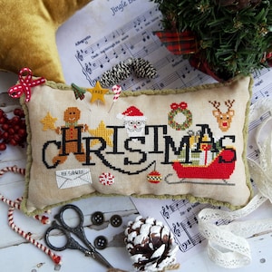 Può includere: Un cuscino beige con un bordo verde presenta un disegno a punto croce della parola "NATALE" in nero con varie icone natalizie, tra cui un uomo di pan di zenzero, Babbo Natale, una ghirlanda, una renna e una slitta con i regali.