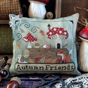 May include: A green and brown cross-stitch pillow with the text "Autumn Friends" and a whimsical scene of mushrooms, a log, a fox, and a bird.