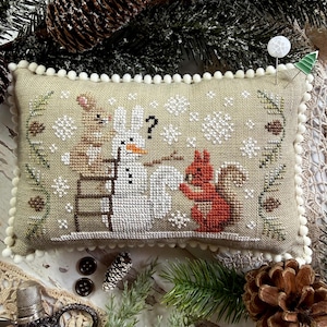 May include: A rectangular beige fabric pincushion featuring a cross-stitch design of a brown rabbit on a ladder, a white snowman with an orange nose, and a red squirrel. The scene includes white snowflakes, pine branches, and a black question mark.