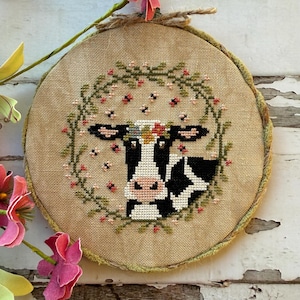 Peut inclure: Broderie au point de croix d'une tête de vache, avec des marques noires et blanches, un nez rose et une couronne florale. La vache est entourée d'une couronne verte avec des fleurs roses et de petits accents noirs. La broderie est dans un cadre rond avec un fond en toile de jute.