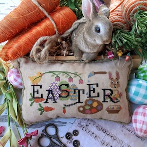 Op de afbeelding: Een beige kussen met een borduurwerkontwerp met het woord "Easter" omringd door kleurrijke eieren, bloemen en een klein konijn. Het kussen is versierd met een bruine franje en ligt op een tafel met andere paasdecoraties.