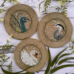 May include: Three circular embroidered pieces with bird designs. A peacock with teal and yellow feathers, a heron, and a white swan are depicted. Each is framed by a green wreath on a tan background.