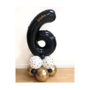 May include: Black number six balloon with the name "Stephen" written in gold lettering. The balloon is on a base of white balloons with gold stars and gold balloons.