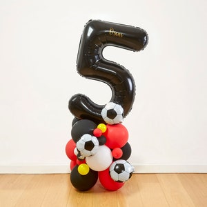 May include: A black number 5 balloon with the name "Oscar" in gold script, atop a cluster of red, black, white, and yellow balloons. Several football balloons are also included in the arrangement. A festive decoration for a birthday.
