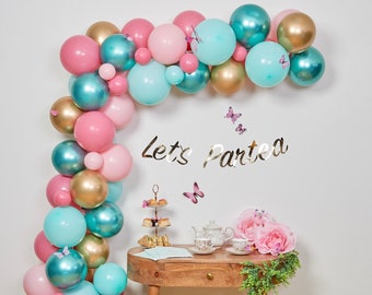 May include: A balloon arch made of pink, teal, and gold balloons with the text "Let's Partea" on the wall behind it. A table with a tea set and a floral arrangement is in front of the arch.