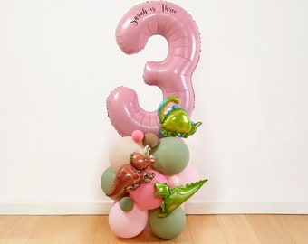 Può includere: Un palloncino rosa a forma di numero tre con la scritta "Sarah is Three". Il palloncino è circondato da palloncini più piccoli di colore rosa, verde e marrone. Ci sono anche piccoli palloncini di dinosauro di colore verde e marrone.
