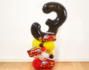 Peut inclure: Ballon noir en forme de chiffre trois avec le texte "Alex is Three" en lettres dorées. Le ballon est entouré de ballons rouges, jaunes et noirs avec des motifs de camions de pompiers.