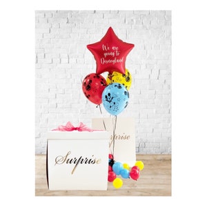 May include: A white box with the word "Surprise" written on it. The box is surrounded by red, yellow and blue balloons with Mickey Mouse designs. One balloon is a red star with the text "We are going to Disneyland" written on it.