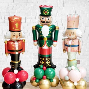 May include: Three colourful nutcracker balloons in red, green, and pink, with gold, red, and green balloon bases. The nutcrackers have detailed faces and uniforms, ideal for festive decorations. The balloons stand tall against a white brick wall.