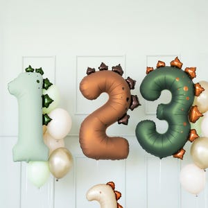Peut inclure: Une scène de fête d'anniversaire avec des ballons en forme de dinosaures et des ballons numérotés. Les chiffres 1, 2 et 3 sont en vert, marron et vert. Deux ballons dinosaures sont en crème et vert. Des ballons blancs, dorés et verts complètent la décoration.