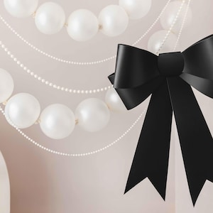 May include: A black satin bow with long tails hangs in front of a garland of white pearl balloons.