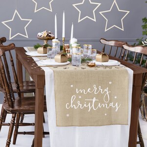May include: A Christmas dining table set with a brown wooden table, chairs, and a burlap table runner. The runner has "merry christmas" in white script. The table is set with plates, glasses, gold-rimmed glasses, and gift boxes. A Christmas tree is in the background.