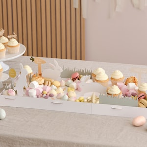 May include: A white tray with a bunny-themed Easter party food display. The tray features white chocolate bunnies, pastel-coloured candies, cupcakes, and macarons. The tray is decorated with green grass and signs that say "This Way" and "Start Here".