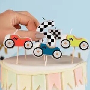 May include: A white cake decorated with a checkered border and race car toppers in red, blue, and yellow. The cake has a banner of pastel flags and a small race car figurine. A hand is placing a checkered flag topper.