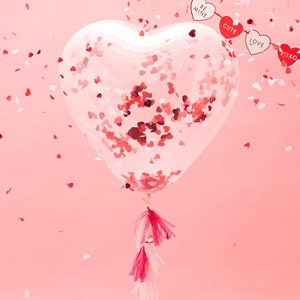May include: A heart-shaped clear balloon filled with red heart-shaped confetti. The balloon has a pink and white tassel and is suspended against a pink background. A banner with the words "BE MINE", "CUTE", "LOVE", and "XOXO" hangs above the balloon.