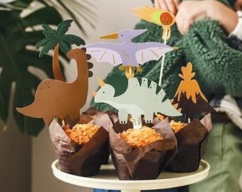 Puede incluir: Un soporte blanco para pasteles contiene cupcakes con decoraciones de dinosaurios. Los adornos incluyen un T-Rex marrón, un estegosaurio verde y un pterodáctilo morado. Dos niños están en el fondo, uno con una camisa a cuadros y el otro con un gorro rojo.
