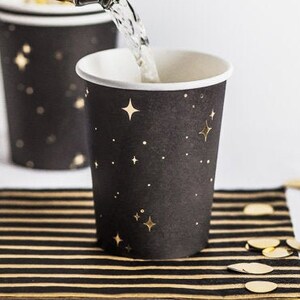 May include: A black paper cup with a white rim and gold star and dot design is being filled with a sparkling beverage. The cup sits on a black and gold striped coaster, with gold confetti scattered around.