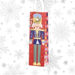 May include: Red wine bottle gift bag with a Nutcracker soldier design. The bag has a white handle and a decorative tag. The Nutcracker wears a blue jacket, red and green striped trousers, and a gold crown. The background is white with snowflake patterns.