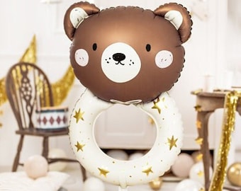 Può includere: Un palloncino a forma di orso marrone con un anello bianco decorato con stelle dorate. L'anello è attaccato alla testa dell'orso e ha uno spago che pende dal fondo.