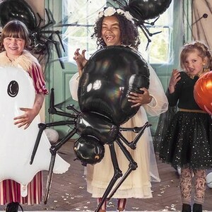 May include: Three children dressed in Halloween costumes hold large inflatable decorations. A girl in a red and white striped dress holds a white ghost balloon. A girl in a white dress holds a black spider balloon. A girl in a black dress holds a pumpkin balloon.